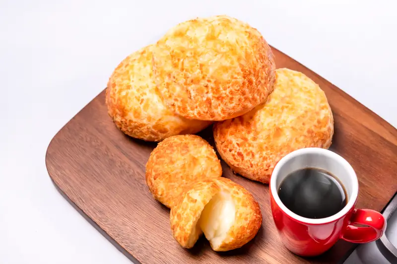 Pães de queijo sobre tábua de madeira, um deles aberto, com xícara de café